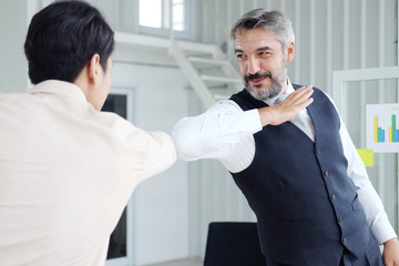 Smiling Senior Caucasian and young Asain businessman greeting with colleagues bumping elbows for preventing covid 19 virus in workspace at office. Social distance and new normal lifestyle concept.