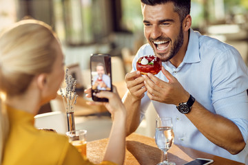 Happy man being photographed by his girlfriend while eating donut in a cafe,