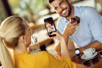 Close-up of woman taking picture of her boyfriend while he's eating donut in a cafe.