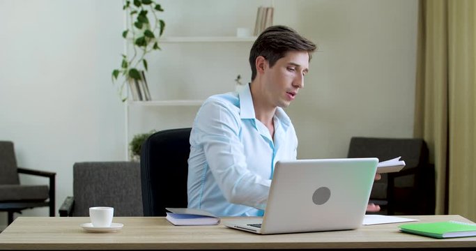 Businessman suffering from summer heat in office home, sitting in stuffy room. Annoyed jaded worker man uses notebooks to freshen himself up with paper documents, air conditioning problems