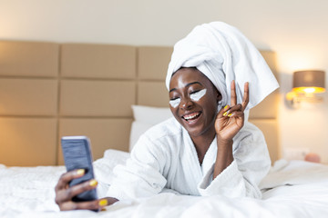 Smiling african woman lying on bed in bathrobe with mobile phone taking a selfie. African woman...