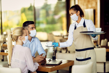 Happy waitress with a face mask serving coffee to a couple in a cafe.