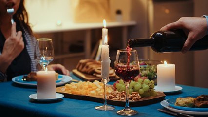 Close up of young man pouring red wine in wife glass. Romantic caucasian happy couple sitting at the table in kitchen celebrating with candle lights, love and anniversary. Romance surprise in relation