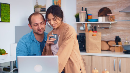 Happy entrepreneur couple working on laptop in kitchen during morning. Husband and wife cooking recipe food. Happy healthy together lifestyle. Family searching for online meal. Health fresh salad