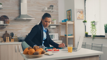 Businesswoman browsing on laptop during breakfast in kitchen. Concentrated business woman in the...
