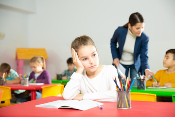 Upset small girl sitting at table in schoolroom on background with pupils studying with teacher