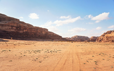 Fototapeta na wymiar Rocky massifs on red sand desert, bright cloudy sky in background, small 4wd vehicle at distance - typical scenery in Wadi Rum, Jordan