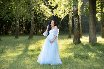 Obraz na płótnie Canvas Pregnant woman posing in a blue dress on a background of green trees.