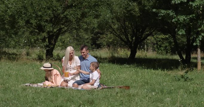 Picnic in nature, parents and children sit on a blanket and drink orange juice, woman pours juice into a glass, family relax in a green clearing.