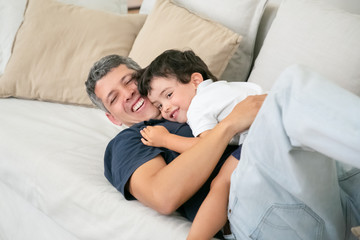 Obraz na płótnie Canvas Smiling father lying on couch with cute little son. Happy handsome Caucasian dad hugging or embracing his lovely child with love on sofa in living room. Fatherhood, family time and parenthood concept