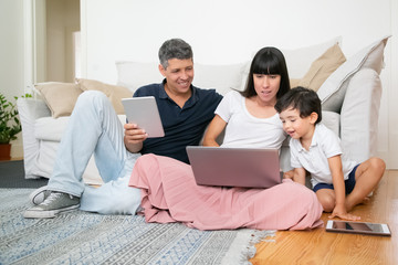 Happy mom, dad and cute little boy using computers, sitting on apartment floor, enjoying leisure time together. Family entertainment and communication concept