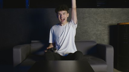 Funny excited young young man gamer winner holding joystick controller playing videogame sitting on sofa feeling overjoyed celebrating victory winning in video game alone at home