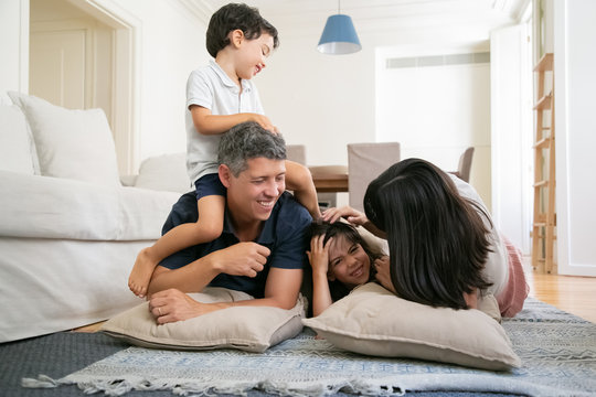 Joyful parents and two little kids having fun together at home, lying on living room floor. Little son sitting on fathers neck, mom cuddling daughter. Family fun time concept