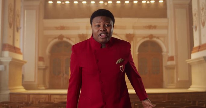Front view of young african american man singing gospel music. Male singer in red suit performing emotionally and moving hands while standing in church. Concept of people and religion
