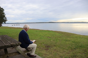 Fototapeta premium Senior male reading a book outdoor overlooking a lake.