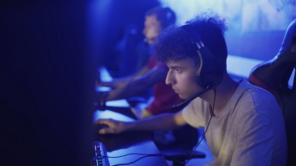 Two friends plays in a computer club, an online game. They are sitting in computer chairs with earphones and playing on the net