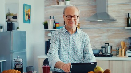 Elderly man surfing on internet using tablet computer during breakfast in kitchen. Elderly person with tablet portable pad PC in retirement age using mobile apps, modern internet online information - Powered by Adobe
