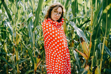 Beautiful caucasian female with short dark wavy hair in glasses wrapped in red and white coverlet enjoys of fresh breathe in cornfield