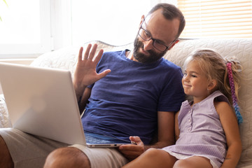 Happy young bearded father with small daughter making video call on laptop computer, gesturing hi, talking with mommy or grandparents using wireless internet connection, sitting on sofa at home