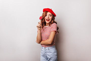 Obraz na płótnie Canvas Blithesome curly woman eating lollipop on white background. Charming french girl in beret posing with candy.
