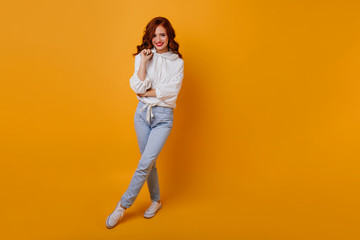 Fototapeta na wymiar Full-length portrait of attractive girl with ginger hair. Indoor shot of happy lady wears white sweater and jeans.