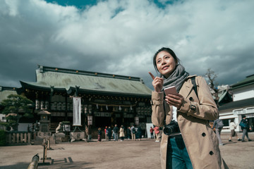 Cheerful beautiful young Asian backpacker blogger woman using smartphone searching tourist attraction location while travel in osaka. happy girl traveler point finger in tenmangu on cloudy day.