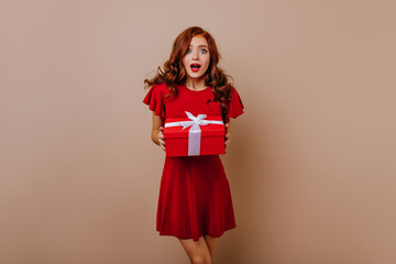 Fototapeta premium Surprised girl in short red dress holding gift. Adorable long-haired woman preparing new year presents.
