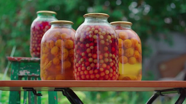 Jars with kompot on table. Sealed glass jars with kompot placed on table near flowers and fruits on summer day in garden