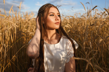 Serene female model in white dress standing in golden wheat field and touching long hair while admiring wonderful sunset in summer in countryside 