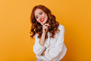 Winsome european girl wears white blouse posing in studio. Indoor photo of elegant red-haired woman smiling on bright background.