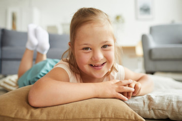 Portrait of little girl with down syndrome smiling at camera while lying on pillows on the floor