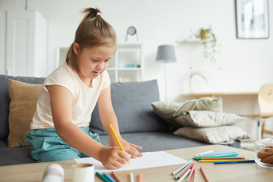 Girl with down syndrome sitting at the table and drawing picture with colorful pencils in the living room at home