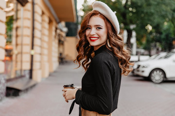 Wonderful ginger woman in beret looking over shoulder. Long-haired smiling girl drinking coffee on the street.