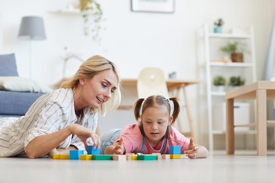 Young mother playing with her daughter with down syndrome in colored blocks while they lying on the floor in the room