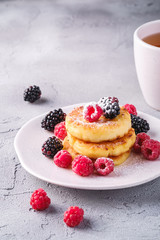 Cottage cheese pancakes and powdered sugar, curd fritters dessert with raspberry and blackberry berries in plate near to hot tea cup with lemon slice on stone concrete background, angle view