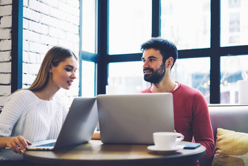 Two male and female freelancers in casual wear having conversation during teamworking on own startup project using wireless internet connection to modern laptop computers sitting in coworking space