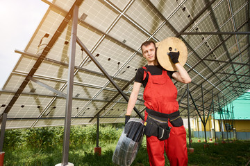 Portrait of man standing under a solar station on a green part near the house with a coil of wires on his shoulder and in his hands. Home construction. Worker in orange uniform and gloves.