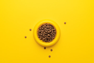 Yellow pet bowl full of dry food on the yellow background. Overhead photo of plastic bowl full of pet food with copy space.