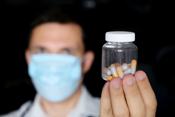 Doctor in medical mask holding bottle of pills, male hand with medication in capsules close up. Concept of pharmacist, drugs, antibiotics or vitamins