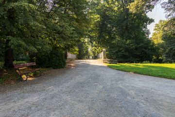 A beautiful bridge to which the road leads. There is a bench by the road. In the background is an alley of a tree.