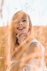 selective focus of excited blonde woman touching neck while looking at camera near grass in meadow