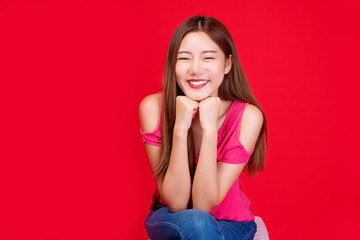 Positive young girl hipster smiling keep her hand under chin while sitting over isolated red background. Friendly woman long hair wear pink t-shirt enjoying in studio.