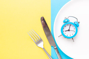 Blue alarm clock, fork, knife and empty plate on colored paper background. Intermittent fasting...