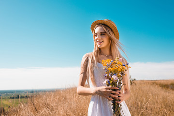 blonde woman in white dress and straw hat holding bouquet of wildflowers while looking away against blue sky
