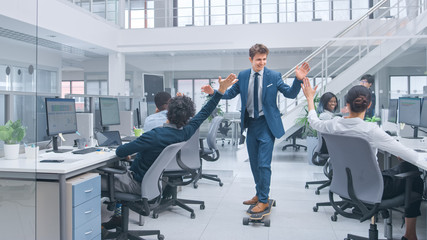 Young Happy Handsome Manager Wearing a Suit and Tie Rides a Longboard. Giving High Fives to Colleagues. Diverse and Motivated Business People Work on Computers in Modern Open Office.