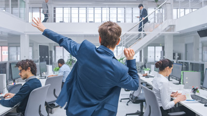 Back View Shot of a Young Cheerful Handsome Business Manager Wearing a Suit and Tie Dancing in the Office. Diverse and Motivated Business People Work on Computers in Modern Open Office.