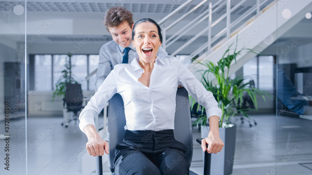 Poster cheerful employee pushes his beautiful female colleague on a chair between rows of desks with divers