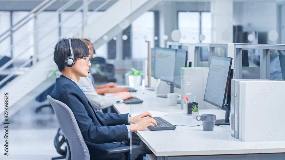 Poster Shot of Handsome and Beautiful Asian Customer Service Operator Working at His Desk Personal Computer in a Busy Modern Call Center. He is Wearing Headsets and Actively Taking Calls. 