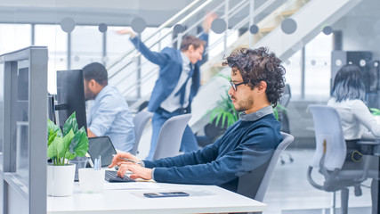 Young Handsome Manager with Curly Hair Works on a Desktop Computer and Compares Data with a Software. Diverse and Motivated Business People Work on Computers in Modern Open Office.