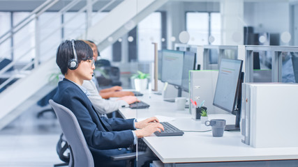 Shot of Handsome and Beautiful Asian Customer Service Operator Working at His Desk Personal Computer in a Busy Modern Call Center. He is Wearing Headsets and Actively Taking Calls. 